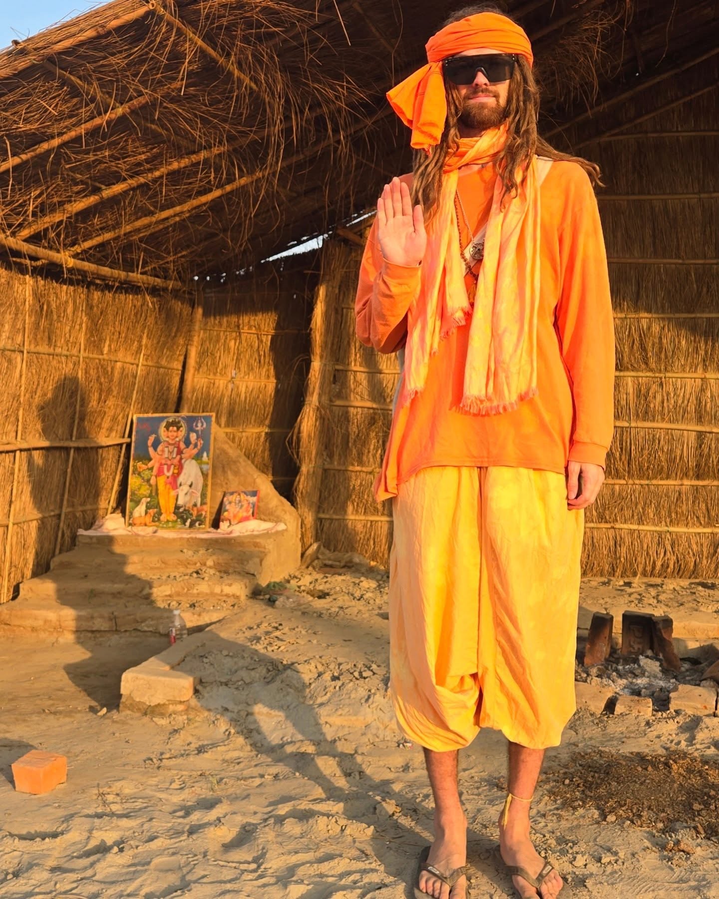 Hunter in full orange sadhu robes and turban, hand raised in blessing before a thatched hut and roadside shrine in India.